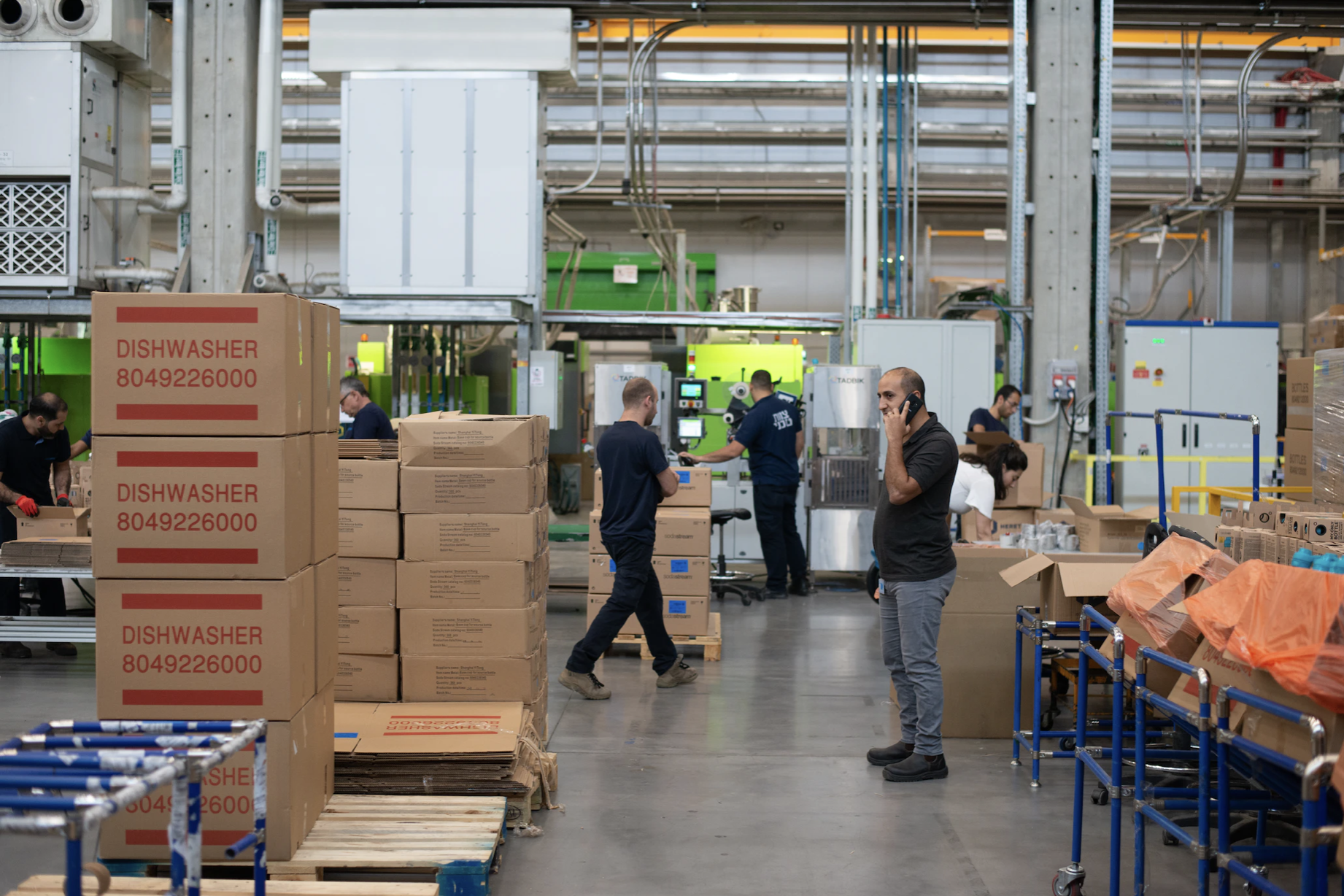 A dishwasher factory floor. Stacks of big boxes labeled "dishwasher." Men moving stacks around. Production line in background.