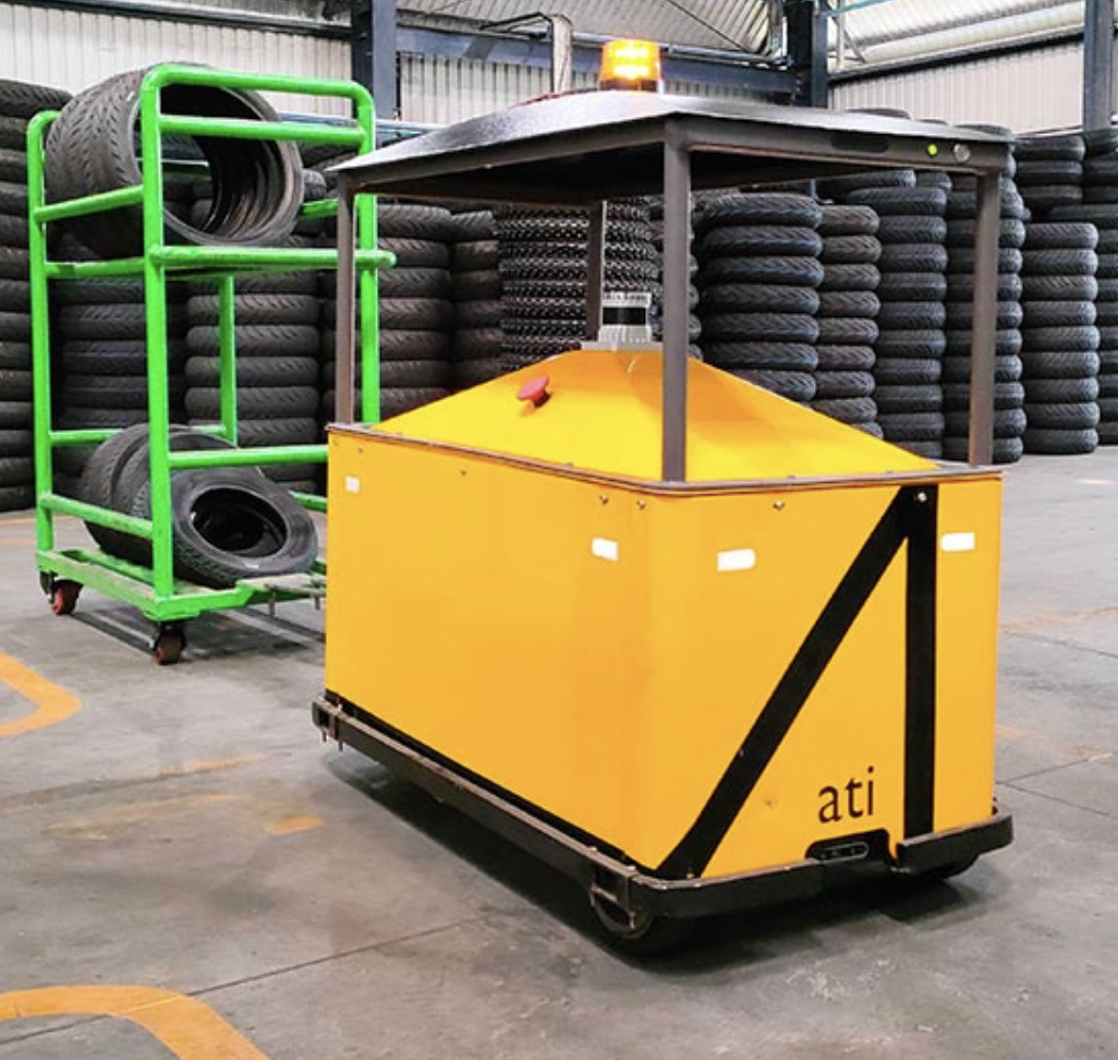 three-wheeled automated vehicle, yellow, 2 feet wide by 2 feet tall by 5 feet long, pulling a green cart carrying motorcycle tires. Stacks of motorcycle tires in the background.